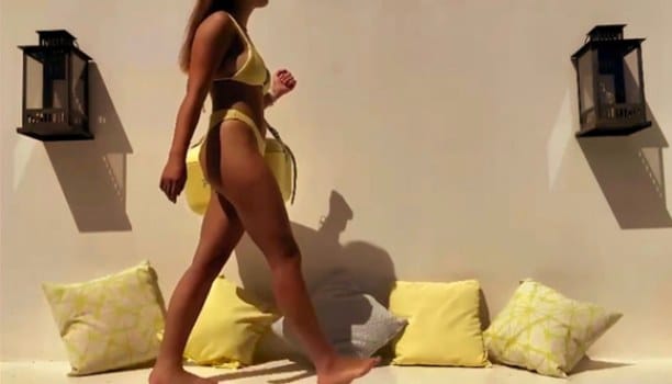 Campaign film still: a woman in a yellow bikini walks past cushions and a white wall in low sun